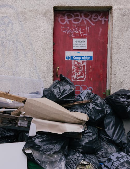 A collection of overflowing rubbish and waste materials piled on a paved urban street corner, including black plastic garbage bags, flattened cardboard boxes, paper and plastic packaging, and loose mi