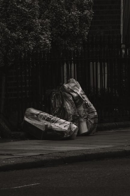 A street scene featuring two large black plastic bags filled with waste, positioned on the pavement near a black metal fence with vertical bars. The bags appear to contain various refuse materials and
